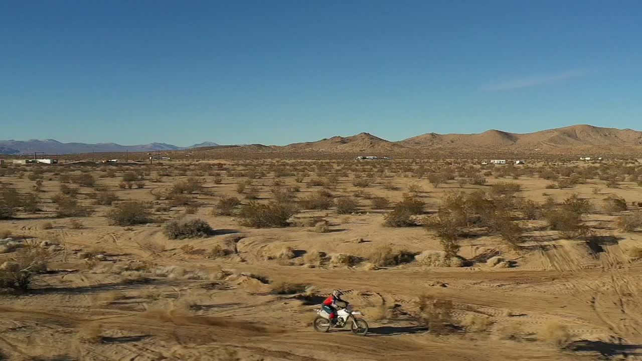 montando una motocicleta a lo largo de un camino de tierra lleno de baches en el desierto de mojave y saltando a través de un camino en cámara lenta - vista aérea