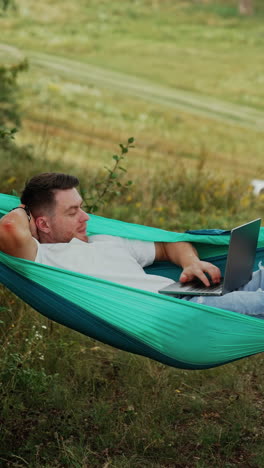 caucasian man lies in the hammock. relaxed freelancer works on laptop remote from the office. vertical video