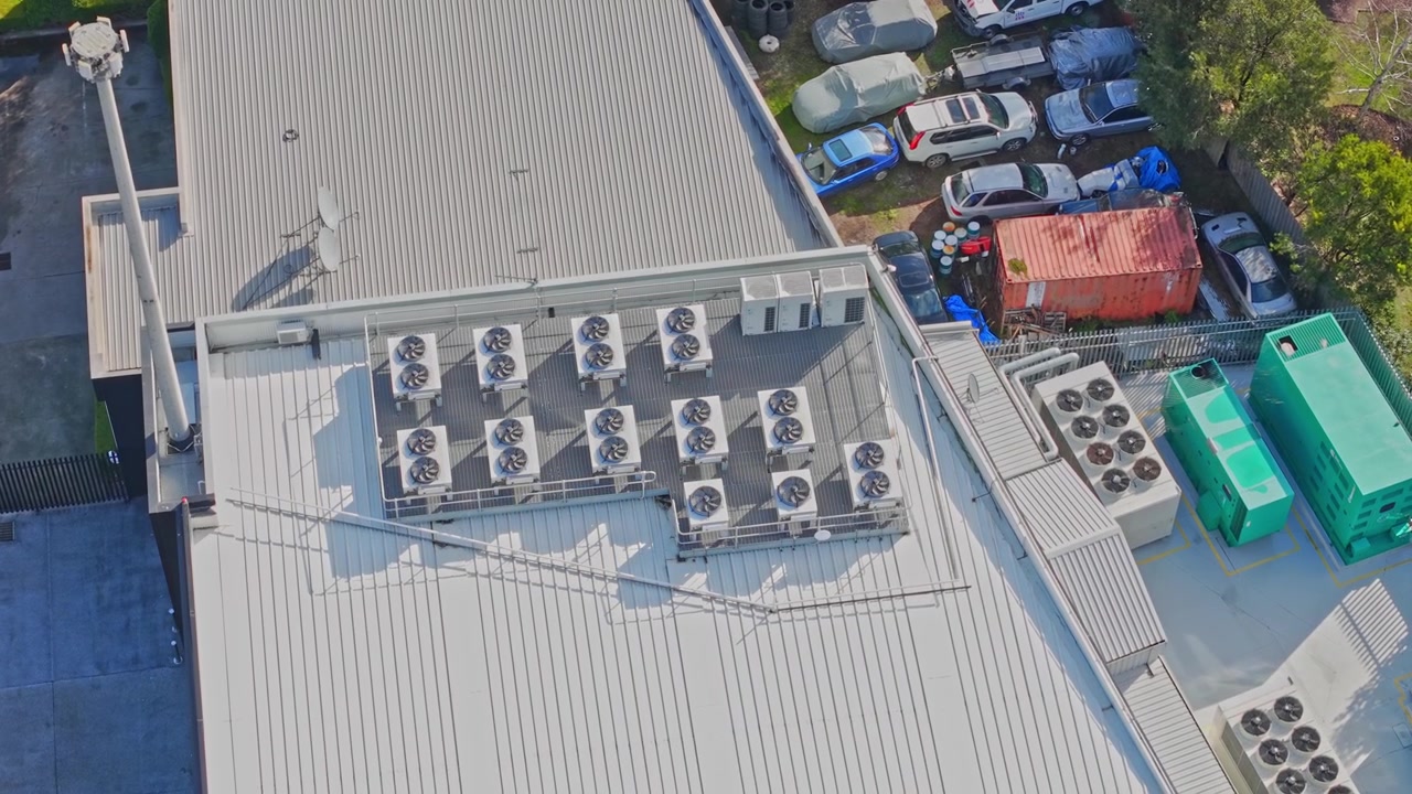 Dynamic aerial footage showcases a modern data centre rooftop lined with industrial HVAC units and large backup generators, surrounded by parked vehicles. Bright natural daylight, sharp clarity.