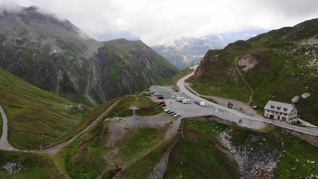 Riprese aeree della sorgente del fiume Reno su un passo montano panoramico con una strada asfaltata, circondato da cime aspre e auto di passaggio, che catturano la bellezza del paesaggio alpino.