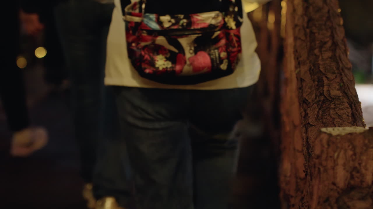 Passeggiata notturna su ponte di legno che mostra la vista posteriore di pedoni con zaino floreale, jeans e sneakers, luci bokeh calde, ringhiere di legno, grana cinematografica e atmosfera cupa della vita notturna urbana.