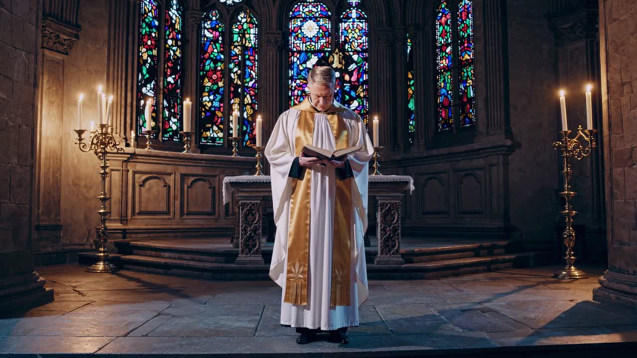 Un sacerdote legge in una chiesa scarsamente illuminata, circondato da vetrate colorate