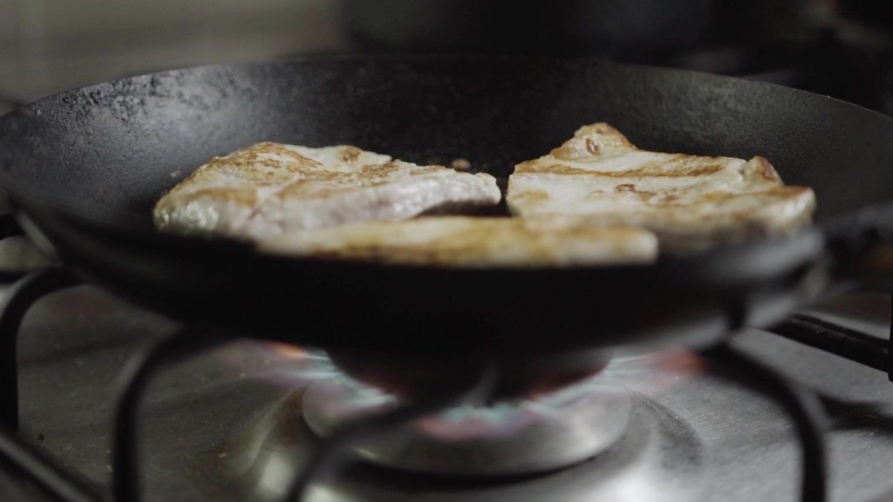 Handheld Slow-Motion Closeup of Pork Chops Being Cooked on Open Flame Gas Stove in Black Cast Iron Skillet