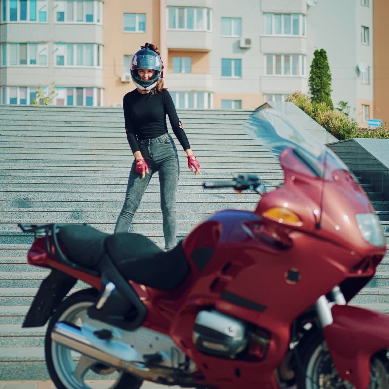 pretty woman in gray jeans and with helmet on her head is posing on the background of a red motorbike and urban high-rise buildings