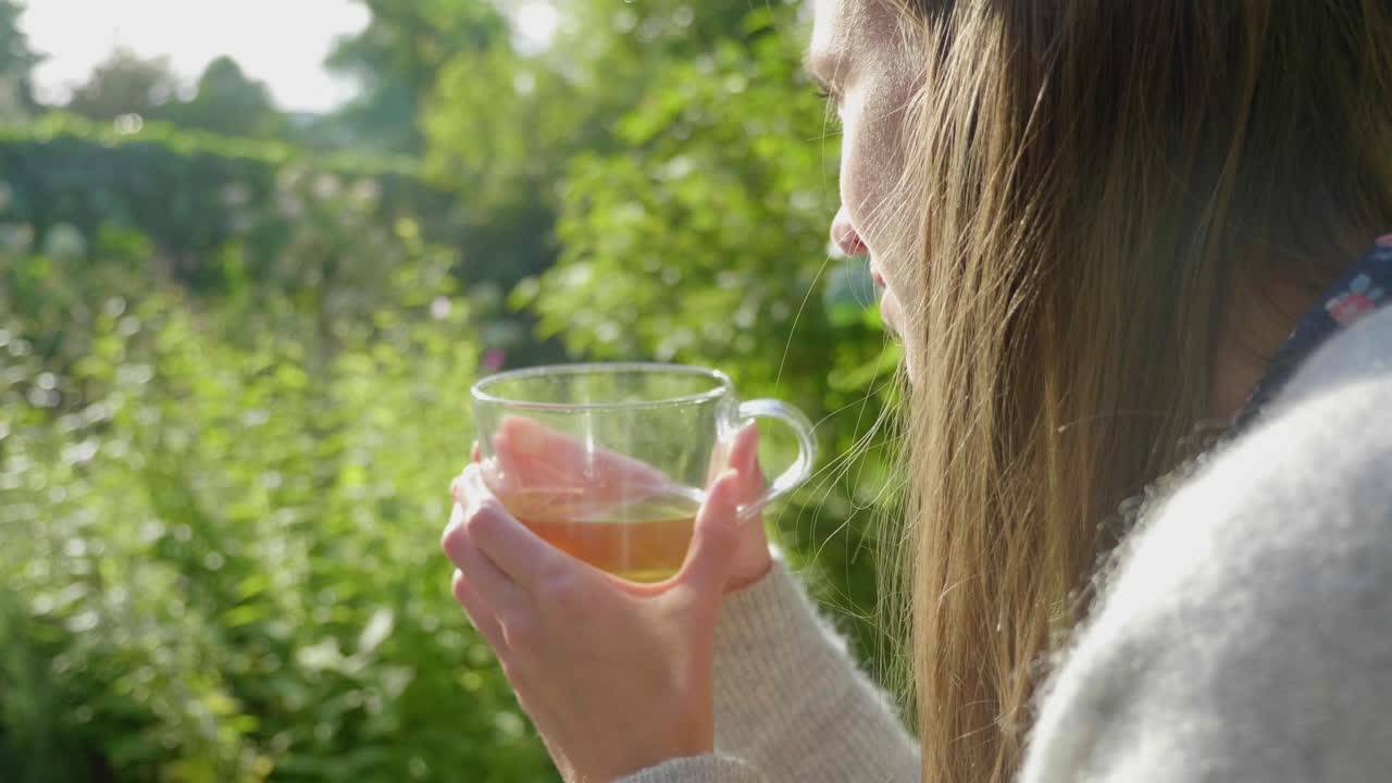 Ragazza con i capelli lunghi sorride con una tazza di tè all'aperto, inquadratura da dietro la spalla.