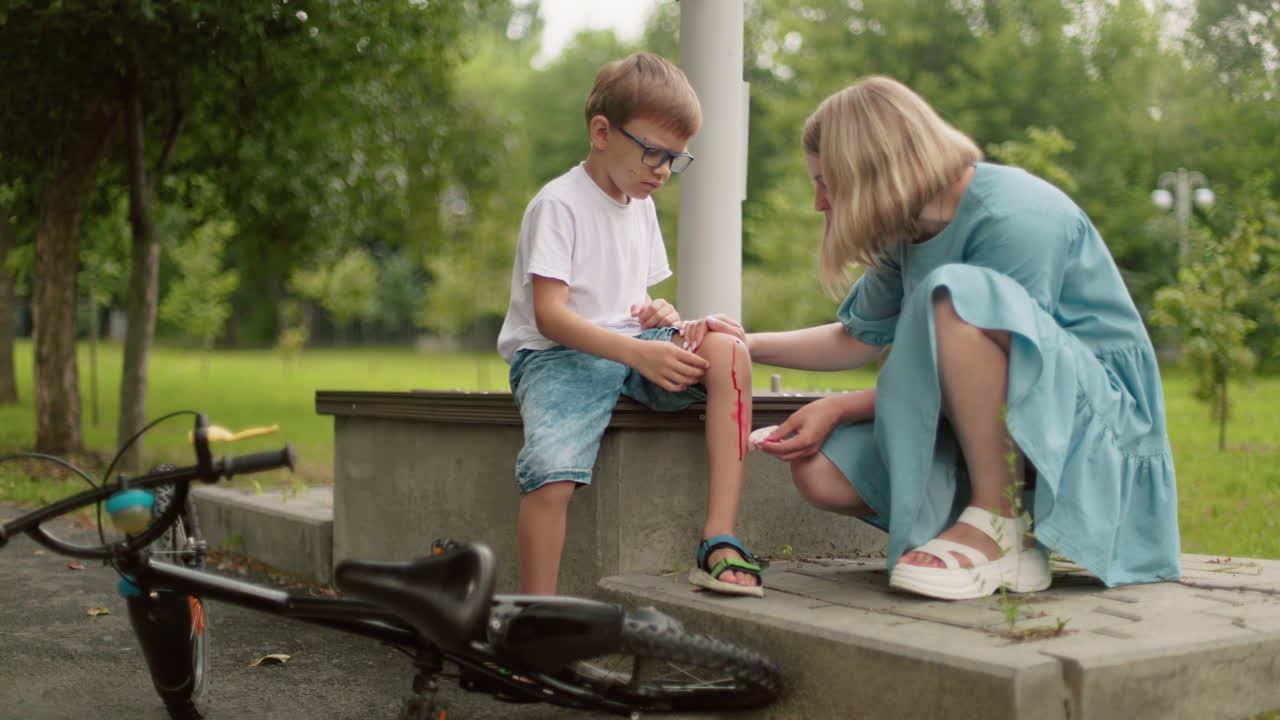 una madre se arrodilla frente a su hijo, limpiando una herida sangrienta en su rodilla con un pañuelo, el niño se sienta en un escalón, mirando infeliz