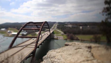 austin pennybacker bridge overlook. focus tilted to lead you off the rock ledge, towards water - isolating the traffic in the center of the arch; bushes on left move wildly in wind.
