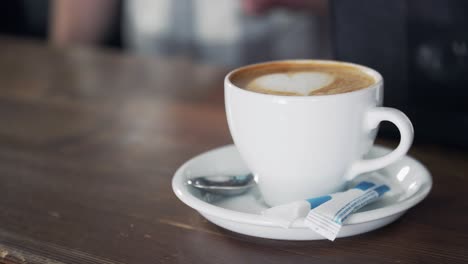 white cup with ready cappuccino standing on wooden table and barman is pouring milk into another cup. delicious drink