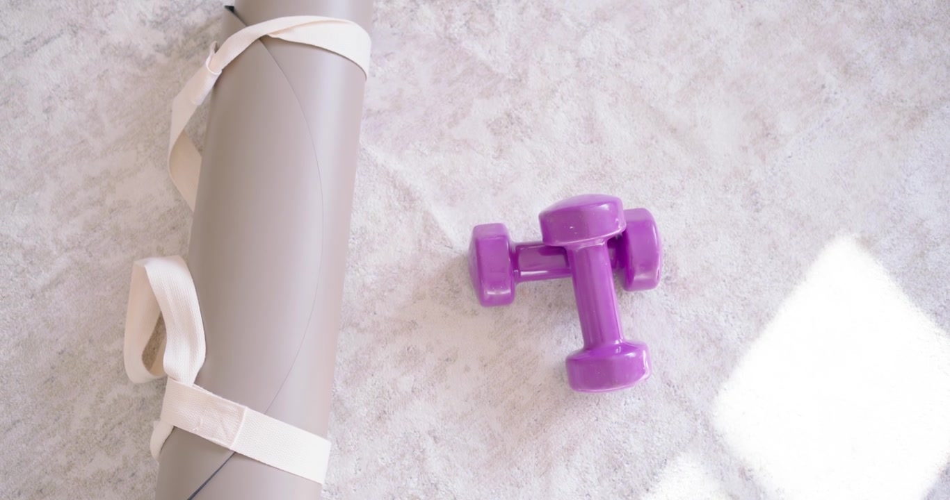 Purple dumbbells are resting on carpet near window as camera is pulling back, revealing rolled mat