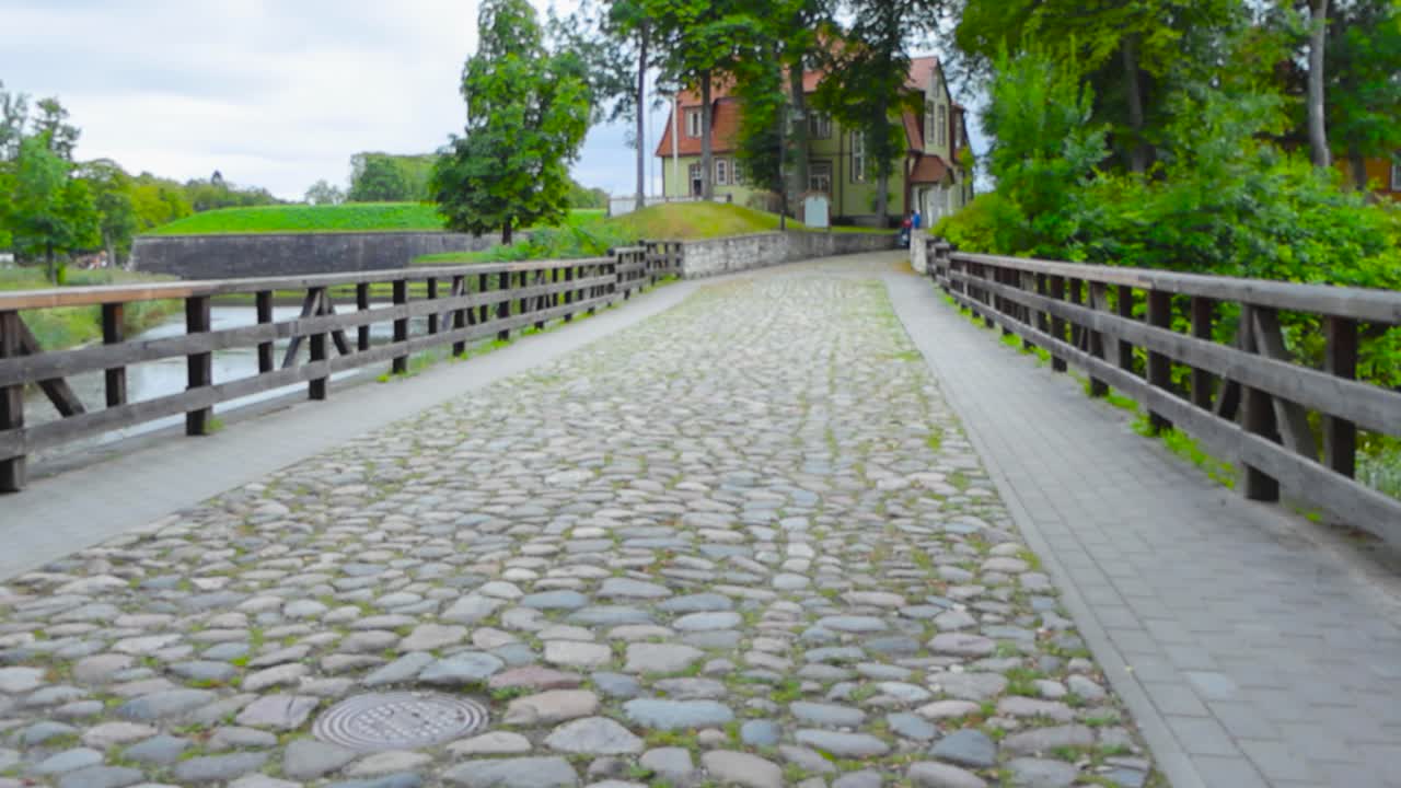 Footage of a cobblestone road during summer day time with moss and grass growing between the rocks. In Kuressaare Estonia the bridge has wooder railings. Bridge goes over a moat or a river.