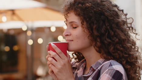 young woman with curly hair smiles while holding a red cup of coffee outdoors