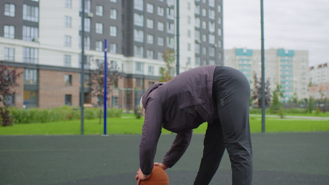 senior man on basketball court warming up, wearing knit beanie and puffer jacket, bending to retrieve ball, urban residential backdrop with apartment blocks and fence, cloudy cool weather, solitary