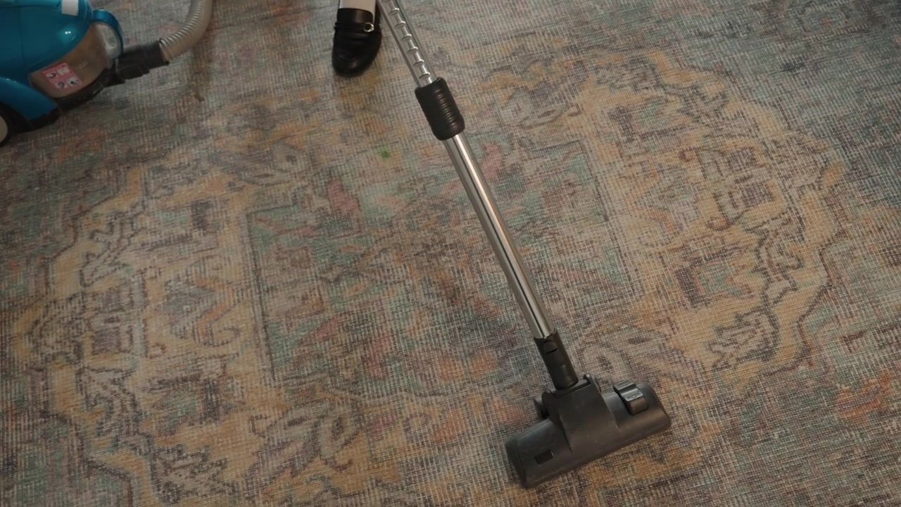 Hlavice vysavače klouzající po vzorovaném koberci, detail úklidu domu ukazující hubici vysavače uhlazující vlákna, jemný pohyb tam a zpět odhalující čistou cestu, texturovaný vintage koberec s teplými tóny, přírodní.