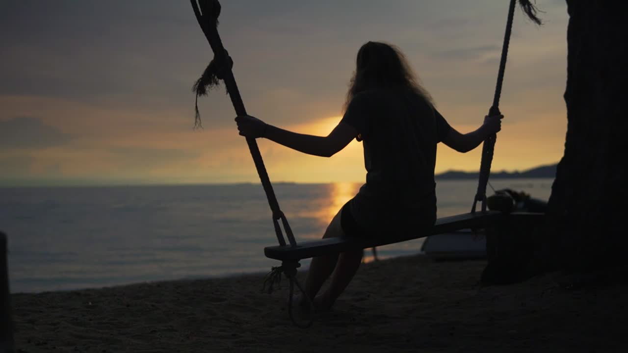 Silhouette di una giovane donna su un'altalena al tramonto su una bellissima spiaggia.