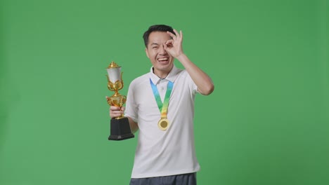 asian man with a gold medal and trophy showing okay hand sign over eye and smiling to camera as the first winner on green screen background in the studio