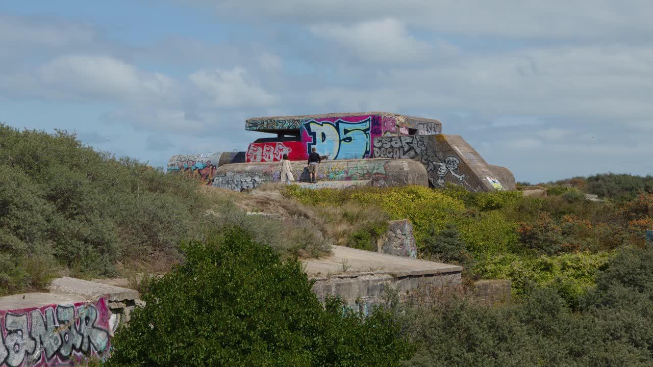 Wide shot of individual walking by abandoned, graffiti-covered concrete bunker under daylight, steady camera