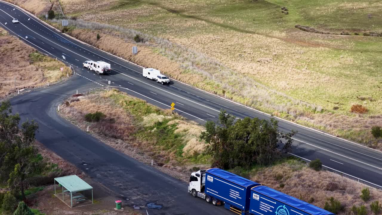 A blue semi-truck and a white caravan travel along a winding rural road in dry, grassy countryside under natural daylight, captured from an elevated perspective