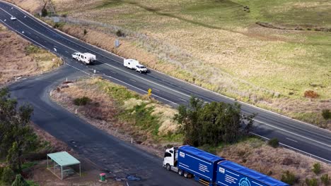 a blue semi-truck and a white caravan travel along a winding rural road in dry, grassy countryside under natural daylight, captured from an elevated perspective