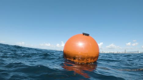 Große-Orangefarbene-Boje,-Die-Ein-Neues-Künstliches-Riff-In-Der-Nähe-Einer-Stadtsilhouette-Markiert,-Bewegt-Sich-Mit-Der-Dünung-Des-Ozeans