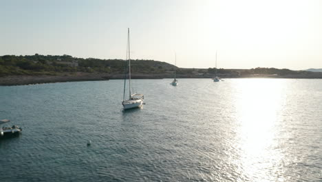 Aerial-forward-view-of-boats-parked-in-a-silent-and-calm-sea-surrounded-by-mountain-and-greenery-at-sunset-in-Ibiza-in-Spain