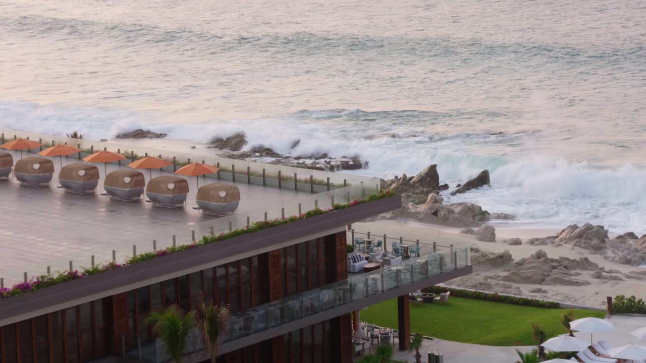 Wide shot of a luxury beach resort with cabanas and waves crashing on a rocky shore, focused on the upscale coastal setting.