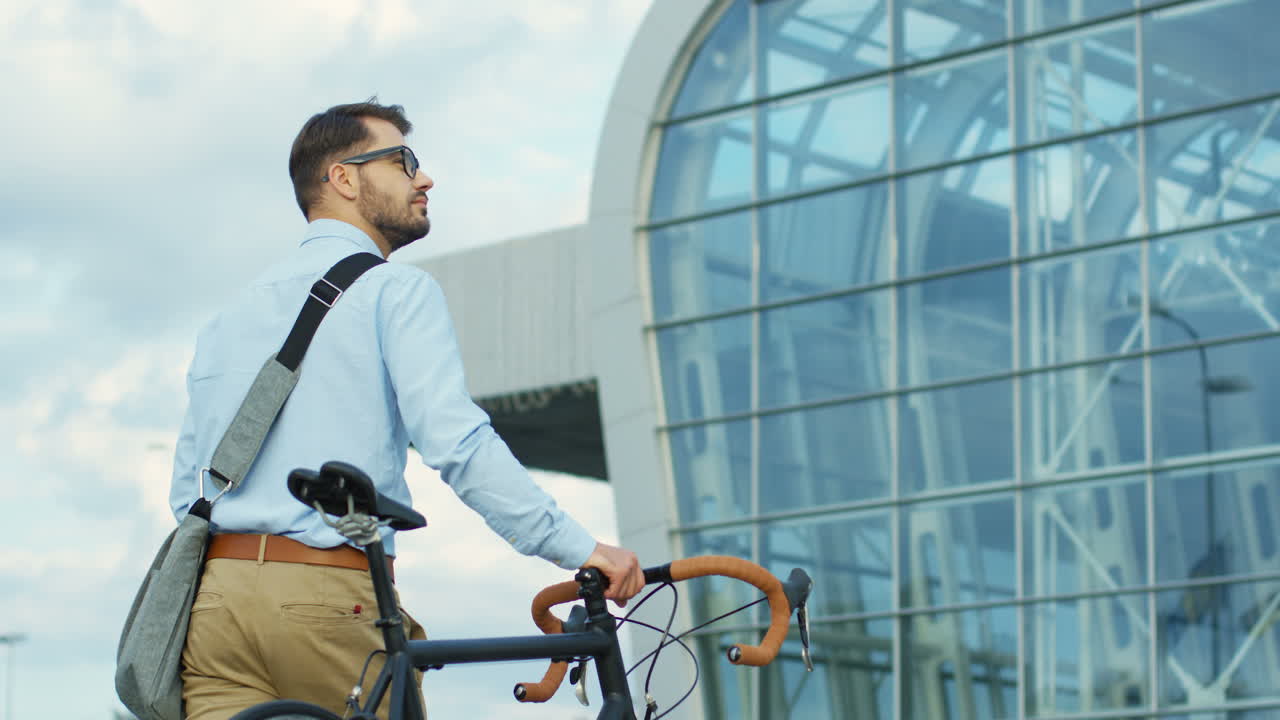 Back view of young man walking to his office building and pulling his bicycle beside him