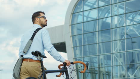 Vista-Trasera-De-Un-Joven-Caminando-Hacia-Su-Edificio-De-Oficinas-Y-Tirando-De-Su-Bicicleta-A-Su-Lado