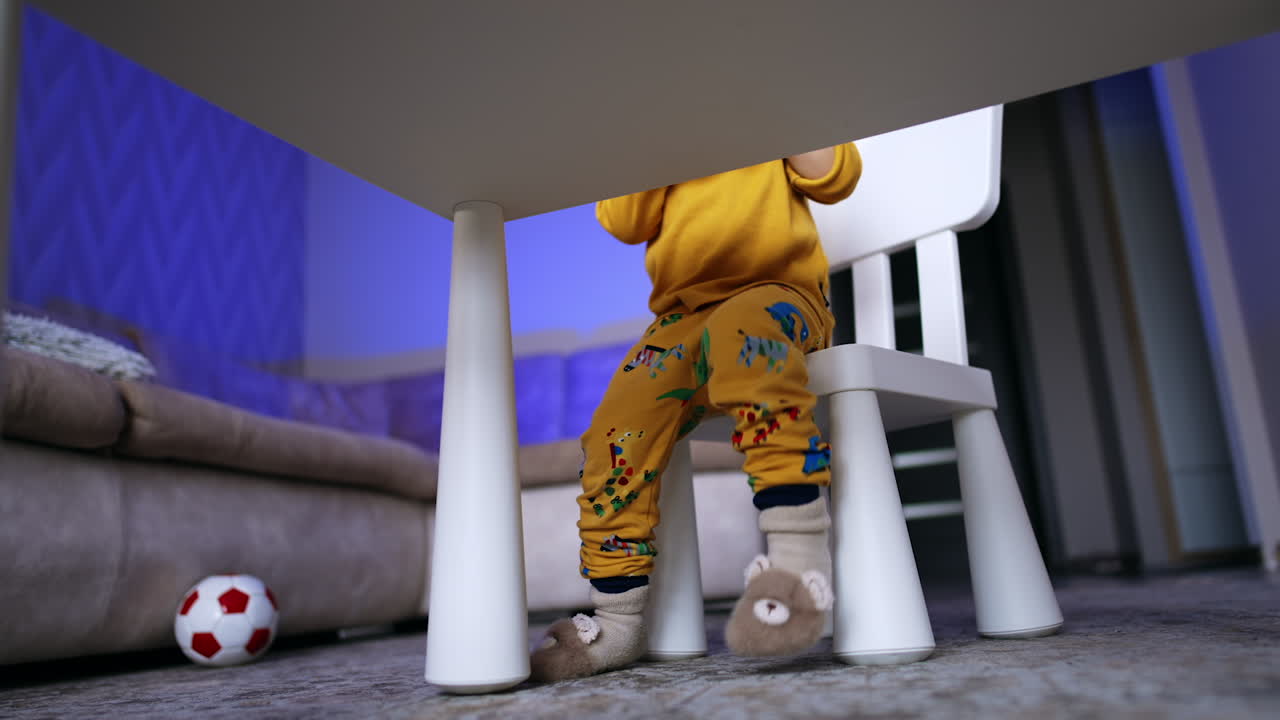 Lower part of a toddler sitting down at the desk. Unrecognized child puts his soft toy on the chair.