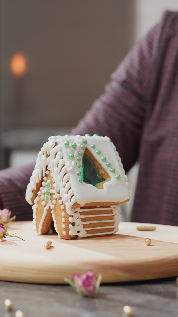 캐터러는 조심스럽게 회전하고 하 아이싱과 녹색 구슬, 나무 트레이, 황금 스프링클, 건조한 꽃, 그리고 배경에 흐릿한 불빛으로 아름답게 장식 된 진저브레드 집을 표시합니다.