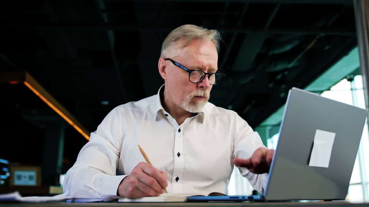 Focused Caucasian entrepreneur works on laptop and takes notes into the paper book. Portrait of a hard-working productive businessman working in office. Low angle view close up.