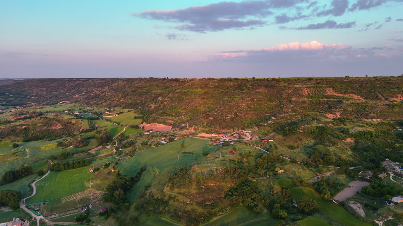 Lateral drone shot flying over agricultural land in La Alcarria, Spain. The scene reveals green hills, scattered farms, dirt roads and warm morning light over the rural landscape at sunrise
