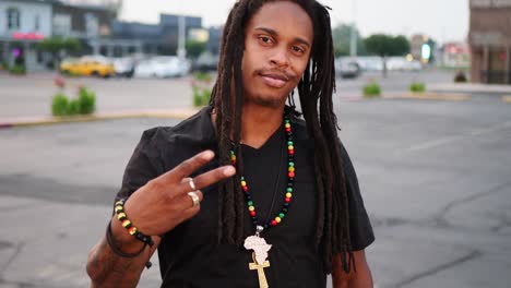 African-American-man-with-dreadlocks-waving-peace-sign,-laughing,-and-being-silly-with-camera-in-open-parking-lot