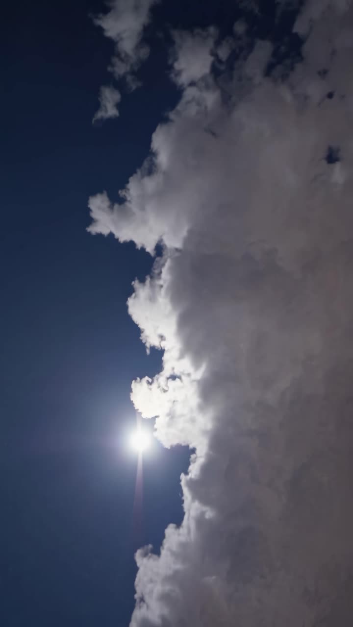 A vertical video captures a dramatic upward angle of a bright sun peeking through thick, fluffy