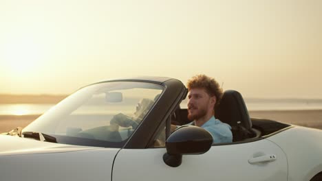 A-bearded-guy-with-curly-hair-in-a-blue-shirt-drives-his-blonde-girlfriend-in-a-white-convertible-car-across-a-field-near-a-river