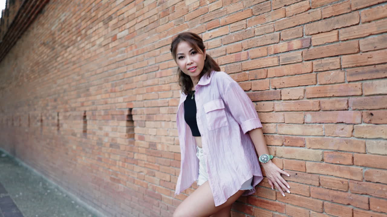 Appoggiata a un muro di mattoni rustico, una donna sorridente con una camicia rosa emana calore e felicità