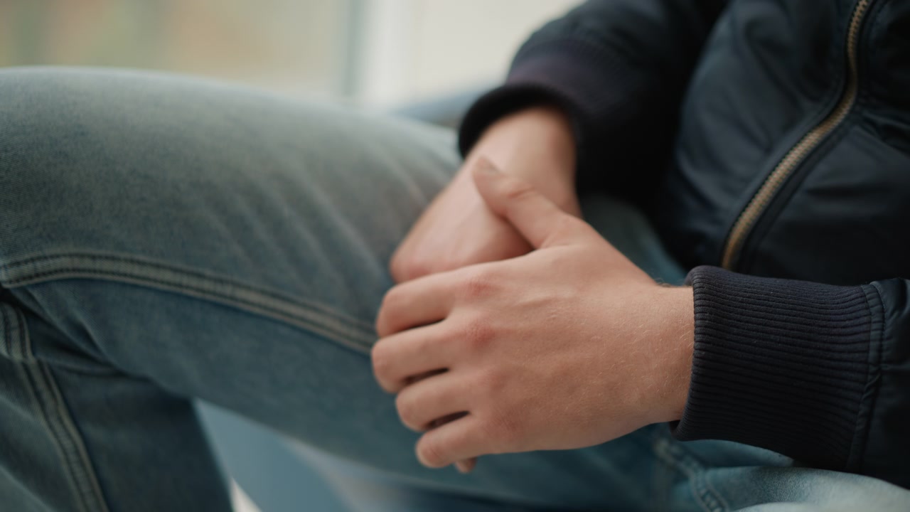 Primo piano delle mani di un uomo appoggiate sulle ginocchia con le dita intrecciate, che indossa jeans blu e una giacca scura. L'illuminazione soffusa evidenzia i dettagli della texture della pelle mentre lo sfondo rimane sfocato, concentrandosi.