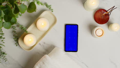 overhead view looking down on still life of blue screen mobile phone lit candles and incense stick with green plant and towels as part of spa day decor