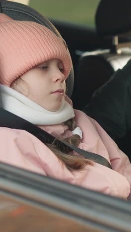 un hombre con una chaqueta verde está desabrochando cuidadosamente el cinturón de seguridad de su hija pequeña, que está sentada tranquilamente en un coche con una jaqueta rosa y una gorra a juego