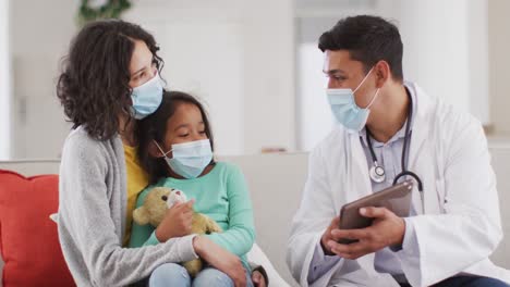 médico masculino hispano hablando con madre e hija en casa, todos con máscaras faciales