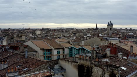 French-city-of-Bordeaux-rooftops-showing-Cailhau-City-Gate-and-pigeon-flocks-flying,-Aerial-pan-right-reveal-shot