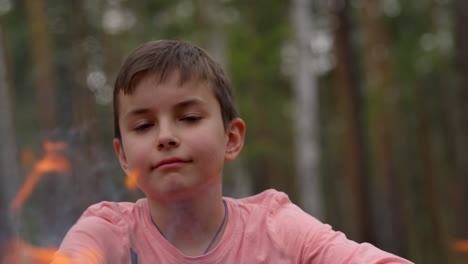 cerca de un adolescente lindo sonriendo a la cámara cerca de una hoguera en el bosque.