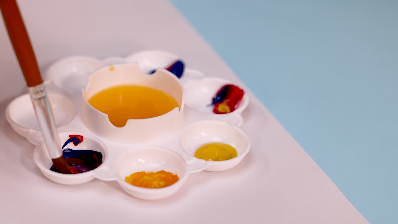 Hand mixes red and blue paint on white palette under soft lighting, close-up, overhead view