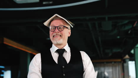 adult male employee in white shirt and black vest holding a paper book on his head. man making faces grimacing in the office.