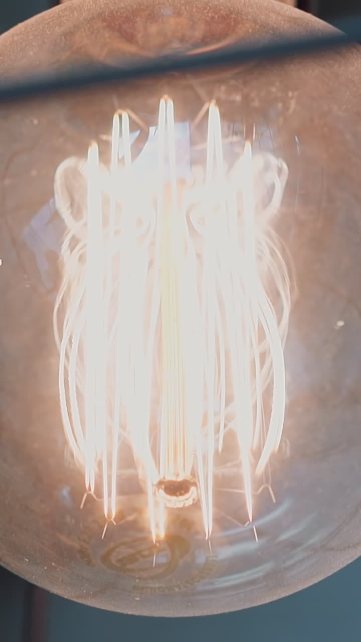 Trembling spiral filament inside glowing light-bulb with stylish metal carcass on ceiling in room macro low angle shot. Contemporary hall interior decor