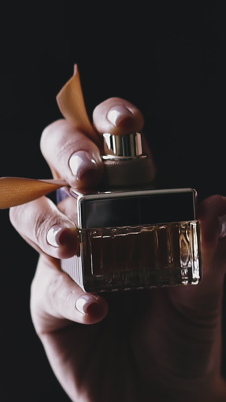 CU, slow motion: young lady in glamorous shiny dress sprays fragrance from square glass bottle decorated with ribbon on neck against black background closeup