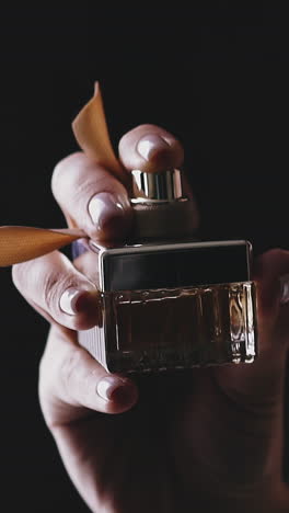 cu, slow motion: young lady in glamorous shiny dress sprays fragrance from square glass bottle decorated with ribbon on neck against black background closeup