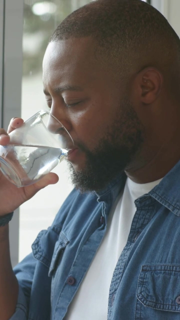 Vertical video: Thirsty man sipping clear glass by window, gazing out in denim shirt white tee band