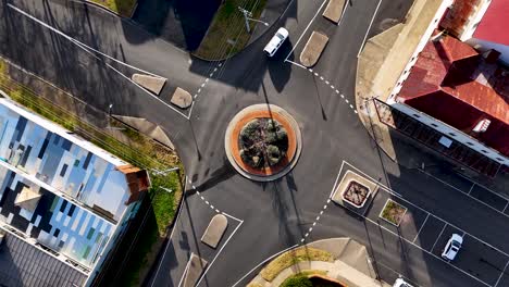 overhead drone footage captures a city roundabout with sparse traffic, commercial buildings, and clear morning sunlight, highlighting urban infrastructure and vehicle movement