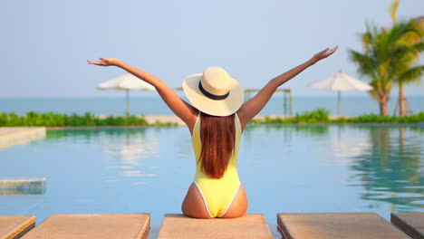 Back-view-of-woman-with-large-hat-and-yellow-swimsuit-sitting-on-pool-edge-and-raising-arms-to-sky