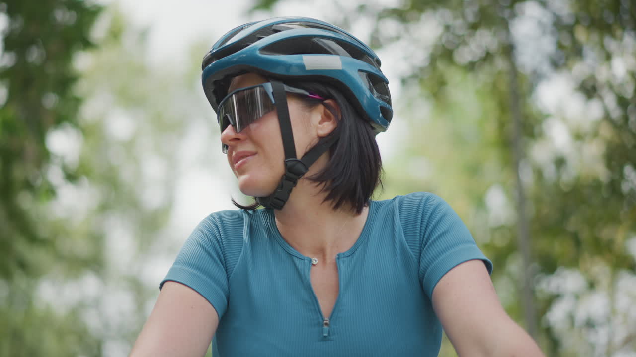 Close up woman cyclist helmet sunglasses smiling toward trees, blue jersey and reflective visor, short dark hair, park background with soft bokeh, confident leisure ride and upbeat outdoor mood